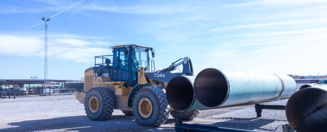 View of tractor loading pipe from a stack onto a truck for transport against a clear blue sky